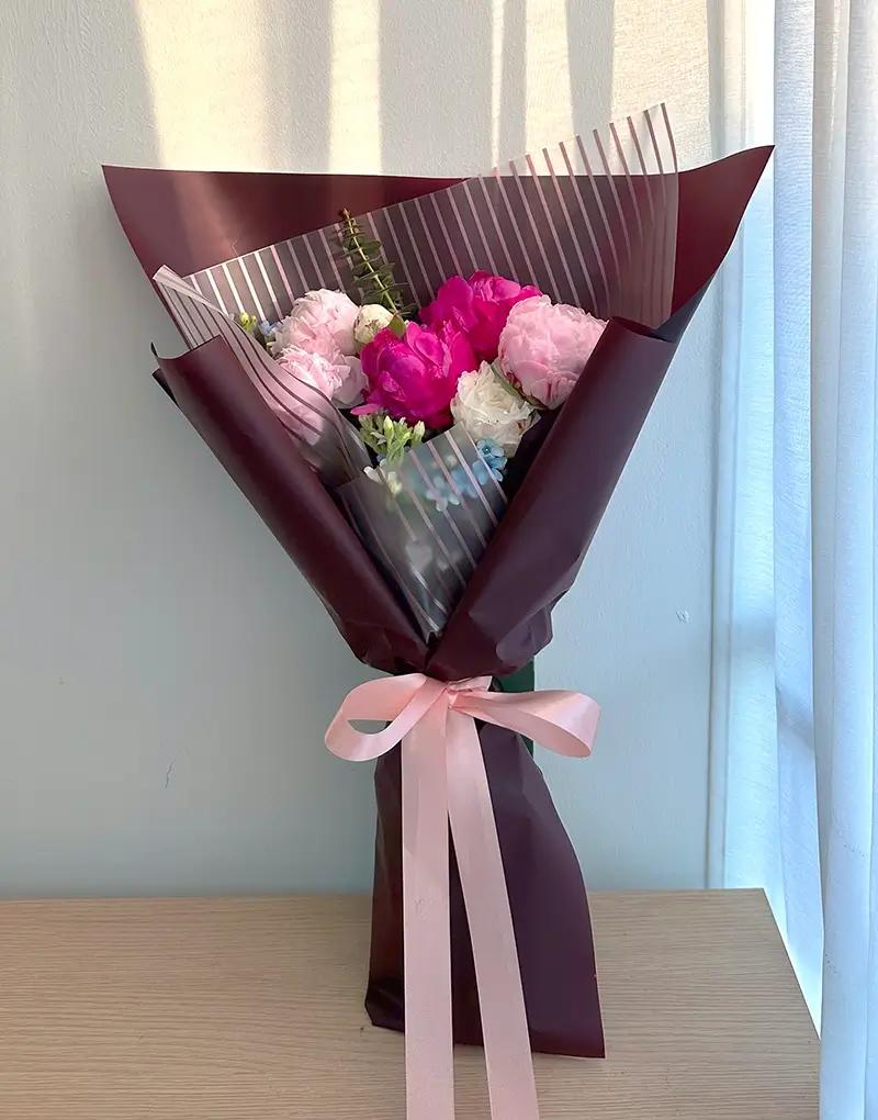 A394 Tall arrangement of dark and light pink peonies with blue and white Oxypetalum, wrapped in maroon paper.
