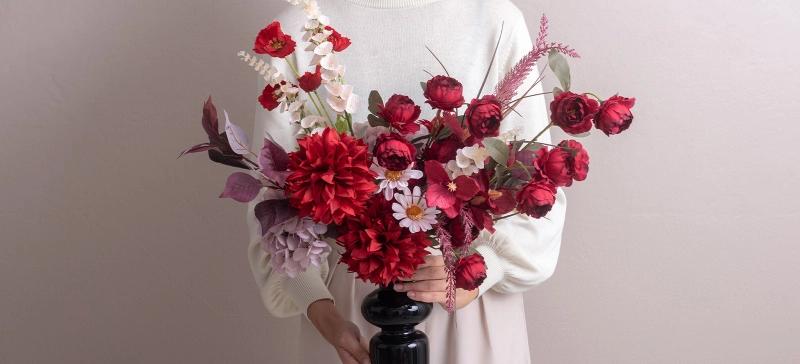 ผู้หญิงในชุดเดรสสีขาว ยืนถือแจกันดอกไม้ประดิษฐ์สีดำ ในแจกันมีดอกไม้ประดิษฐ์สีแดง