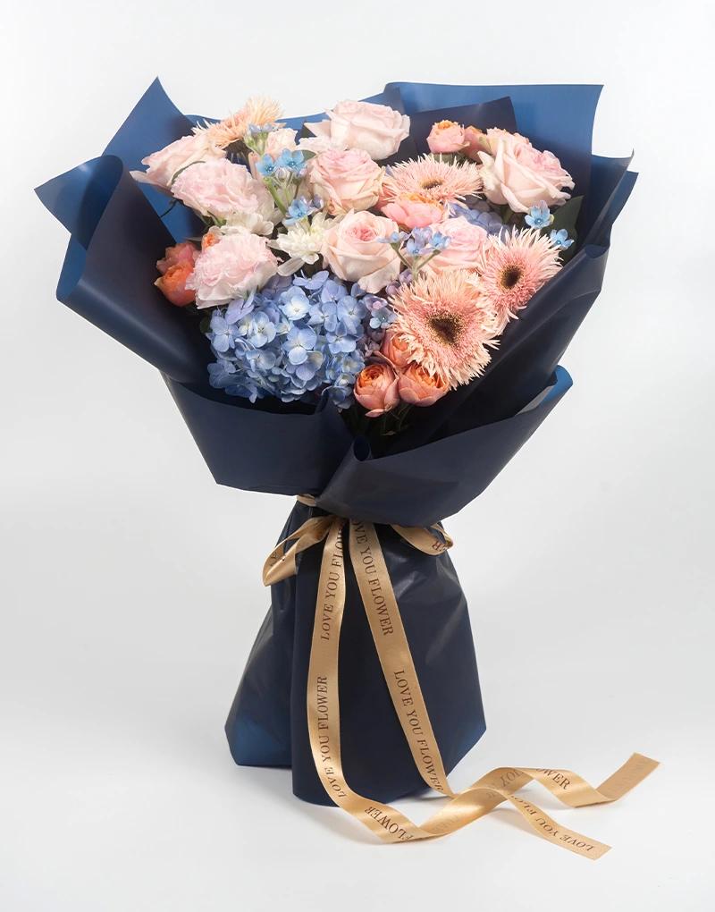 ช่อดอกไม้สดโทนสีชมพู พีช ฟ้า และขาว ห่อกระดาษสีน้ำเงินเข้มผูกริบบิ้นสีทอง