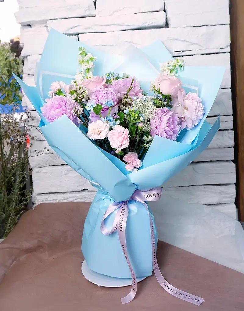 ช่อดอกไม้สดโทนสีชมพู ม่วง และขาว ห่อกระดาษสีฟ้าอ่อน
